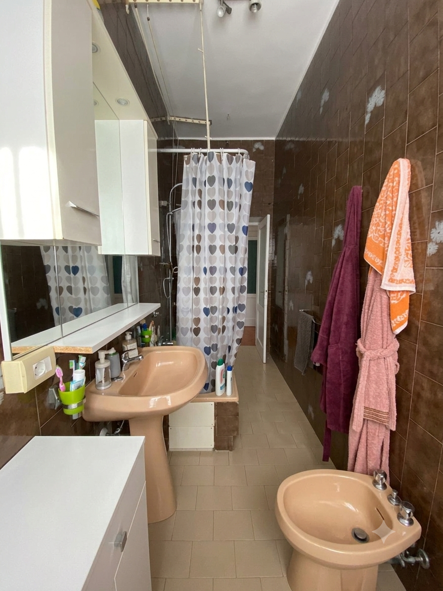 Una spaziosa stanza da bagno con pareti rivestite di piastrelle marroni. Al centro c'è un lavandino color crema con vari saponi e spazzolini, sopra uno specchio con armadietti. Sul lato destro, due asciugamani appesi, uno viola e uno rosa, e un bidet