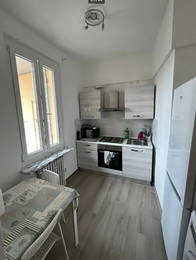 Cucina compatta con mobili chiari, piano cottura a gas, forno, lavabo, frigorifero bianco, tavolo con sedie, e vari oggetti da cucina vicino alla finestra.