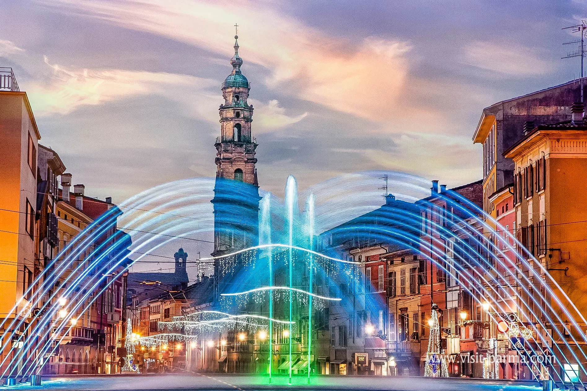 Una piazza con fontane illuminate con luci di diversi colori, con edifici storici colorati ai lati e un campanile in stile barocco sullo sfondo, durante il tramonto o al crepuscolo.