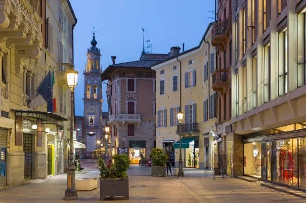 Via pedonale in centro città con edifici storici e negozi, illuminata da lampioni serali.
