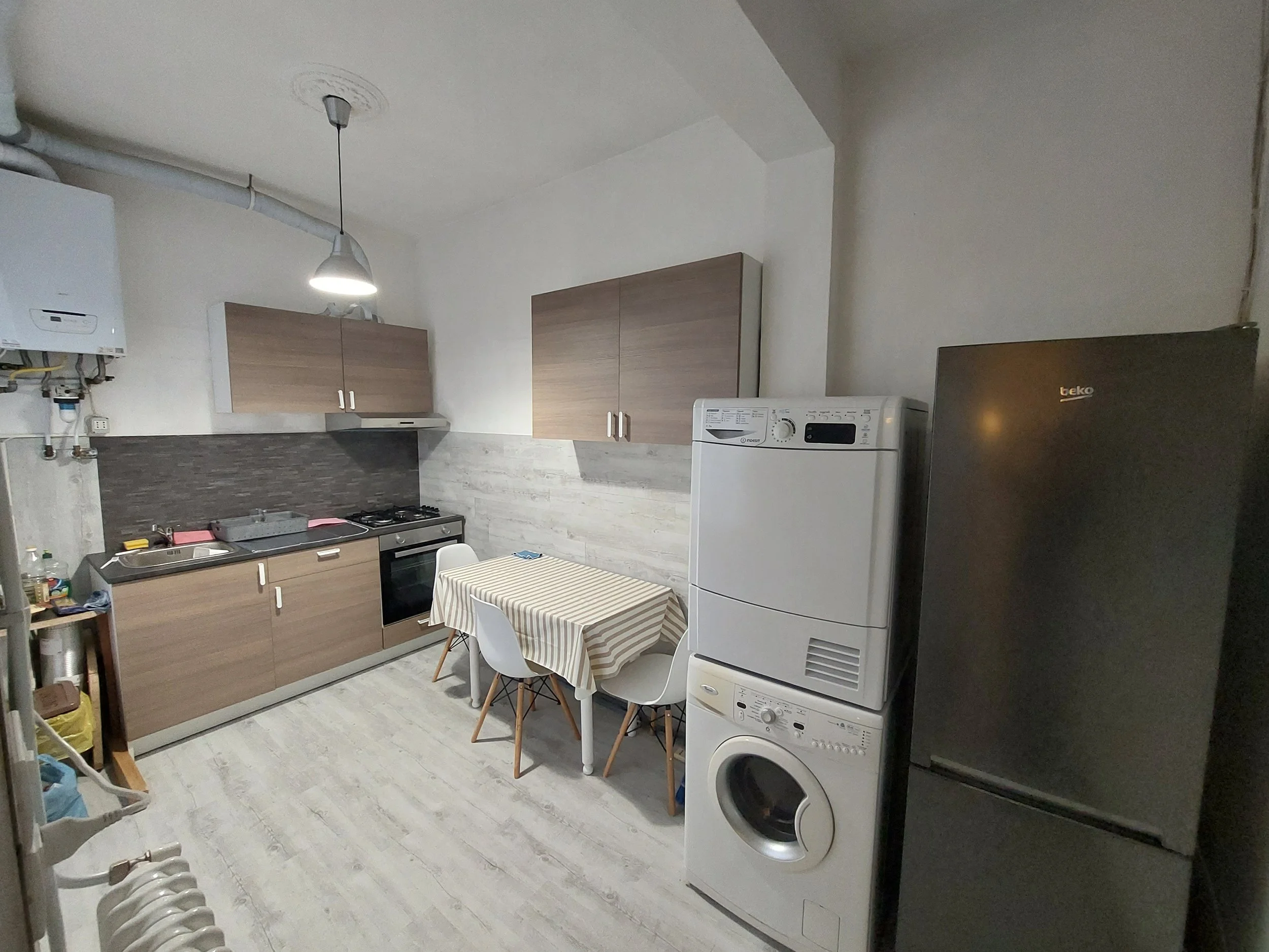 Interno di una cucina con mobili in legno chiaro, una lavatrice, e un frigorifero nero. Ci sono tavolo con tovaglia a righe e due sedie, mentre sul soffitto si vede una luce sospesa.