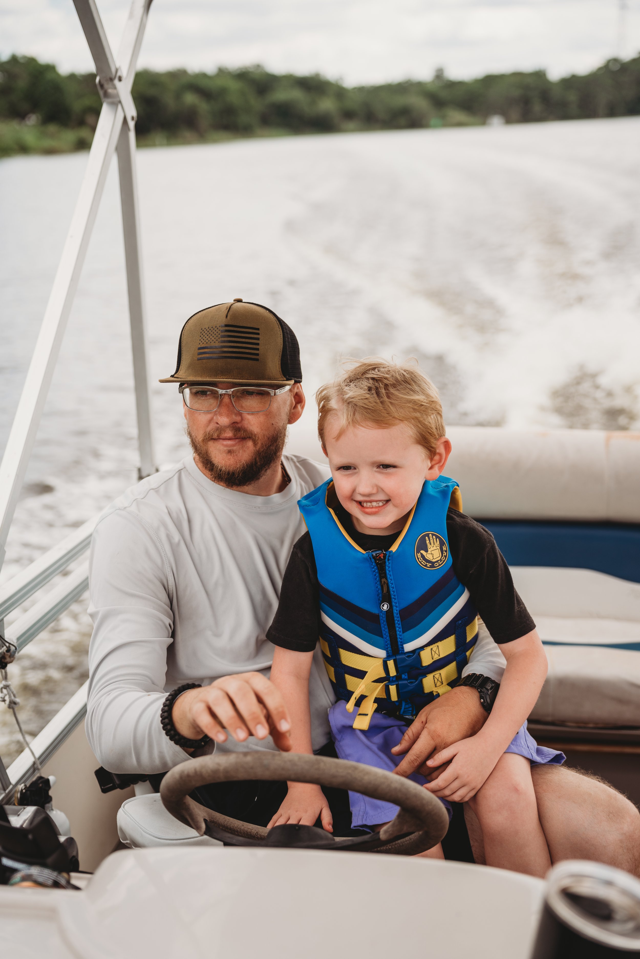 A man with a beard and glasses wearing a baseball cap and a white long-sleeve shirt, and a young boy with blond hair in a black life jacket, sitting on a boat with water and greenery in the background.