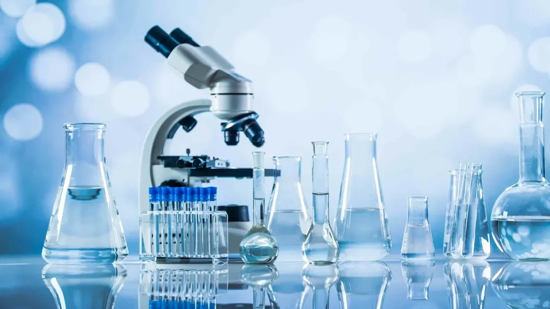 Laboratory scene with a microscope, test tubes, beakers, and flasks filled with clear liquids on a reflective surface.