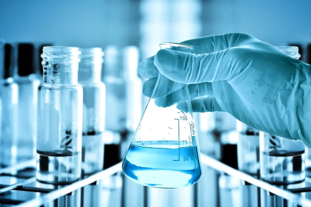 A scientist wearing gloves holding a laboratory flask filled with a blue liquid, with test tubes in the background.
