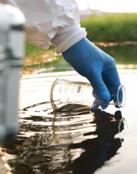 Person wearing blue glove and white protective suit pouring liquid from a beaker into water.
