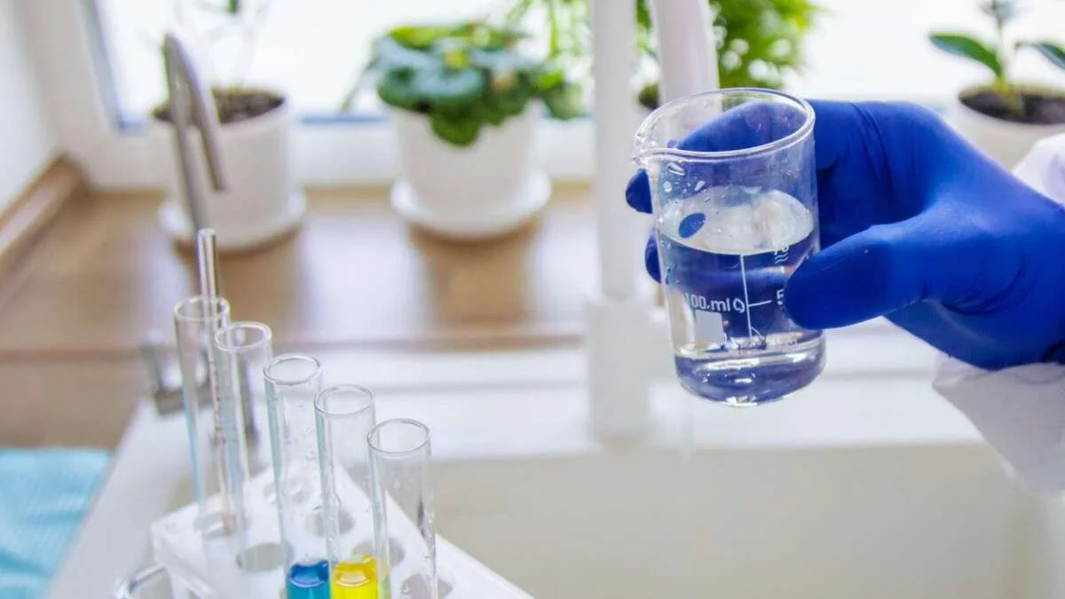 A person in a blue glove holding a beaker with clear liquid in a laboratory.