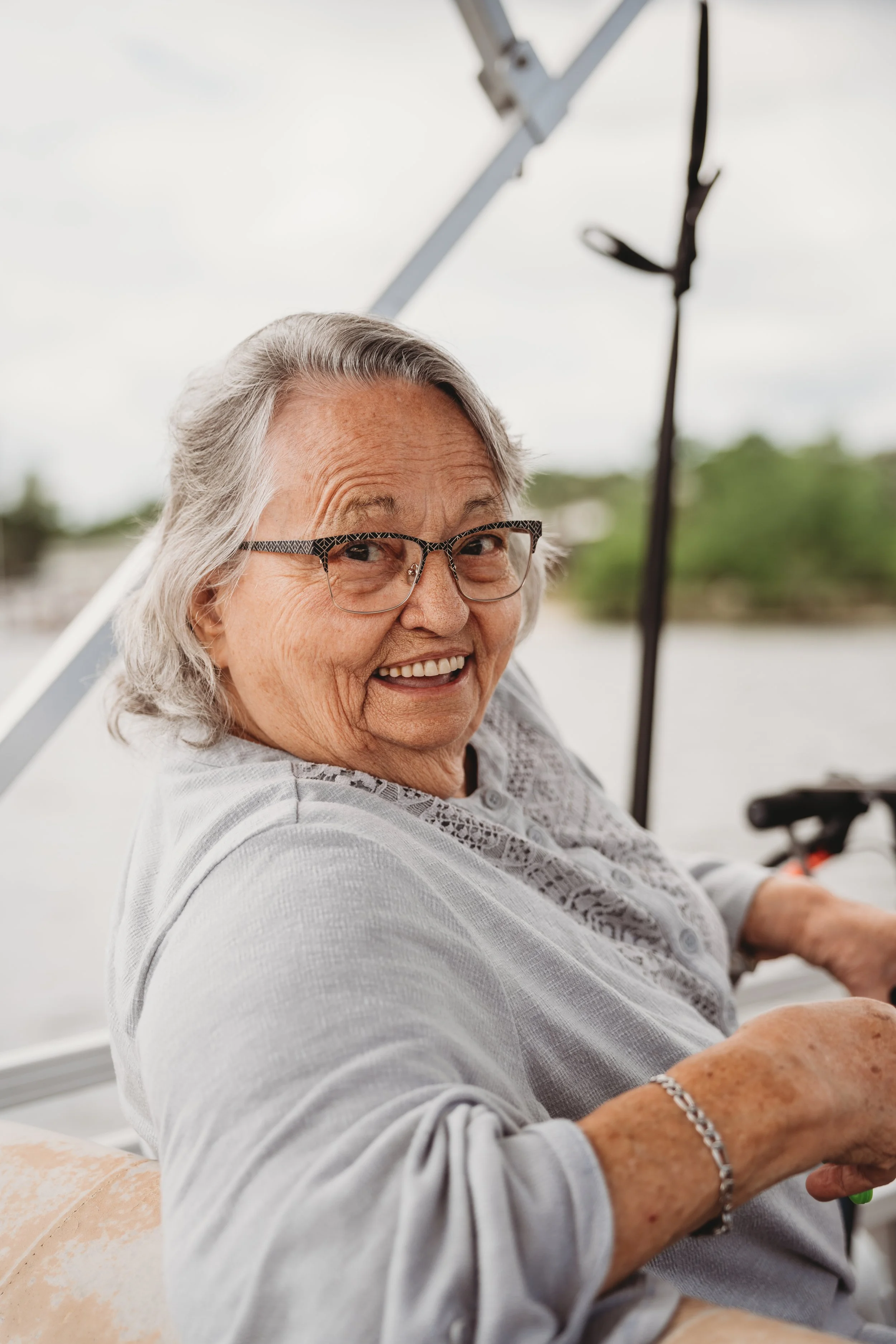 An elderly woman with gray hair and glasses smiling while sitting outdoors in a wheelchair.