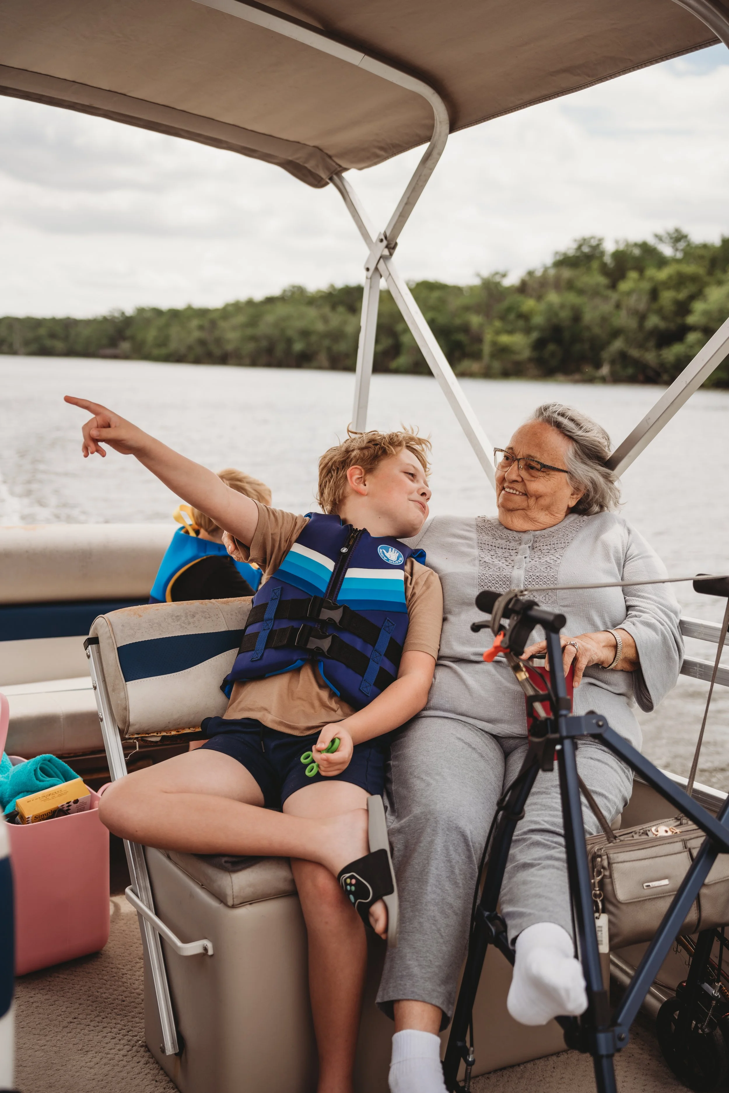 A smiling elderly woman sitting on a boat with a young boy, pointing at something in the distance. The boat is on a river with trees on the shoreline and a cloudy sky overhead.
