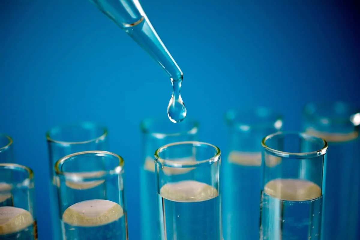 A laboratory scene with multiple test tubes filled with a yellowish liquid and a dropper releasing a droplet into one of the test tubes against a blue background.