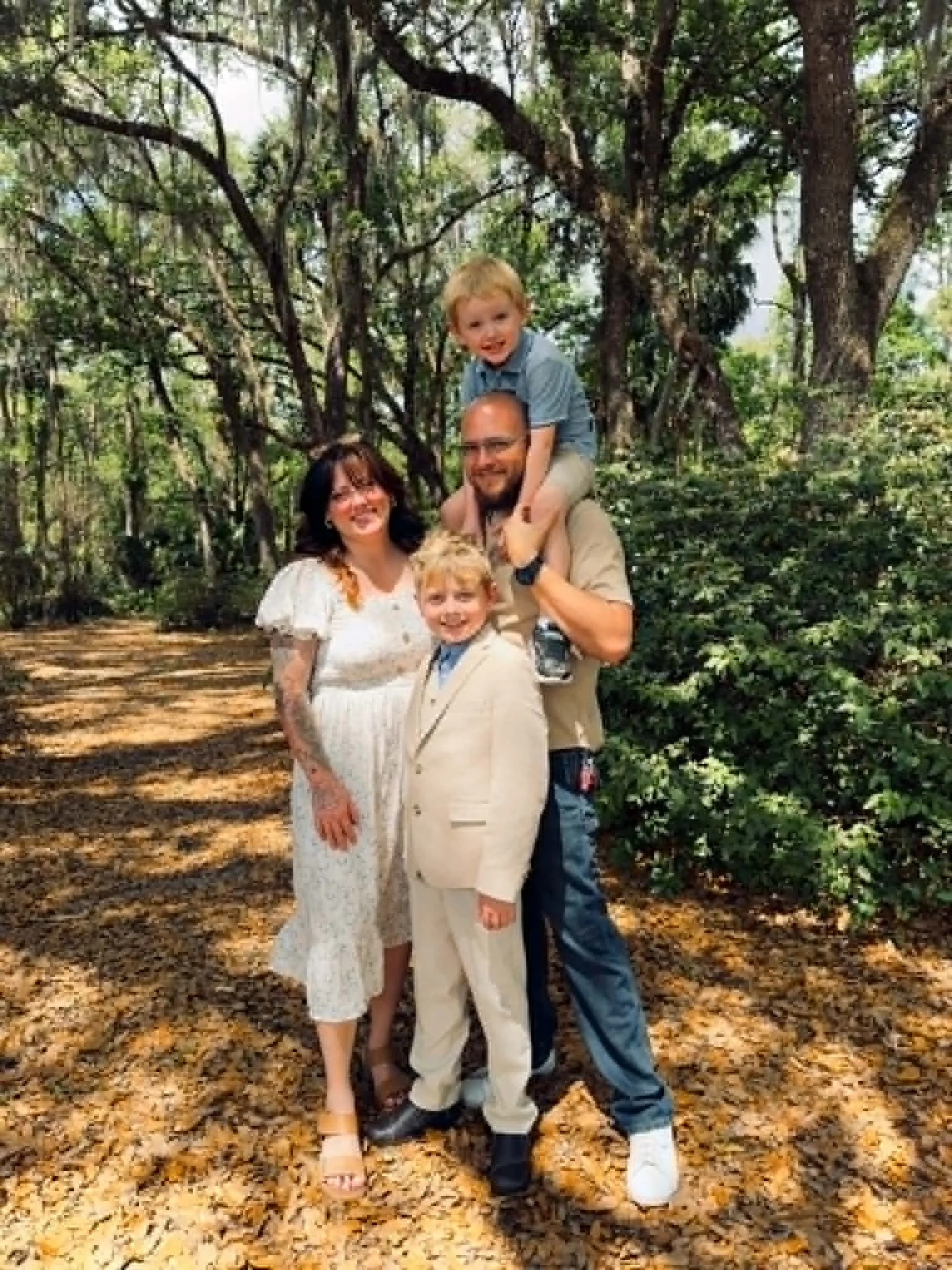 Family of four standing on a forest path with fallen leaves, surrounded by trees and greenery. The man is carrying a young boy on his shoulders, and a woman and a young girl stand beside him, all smiling.