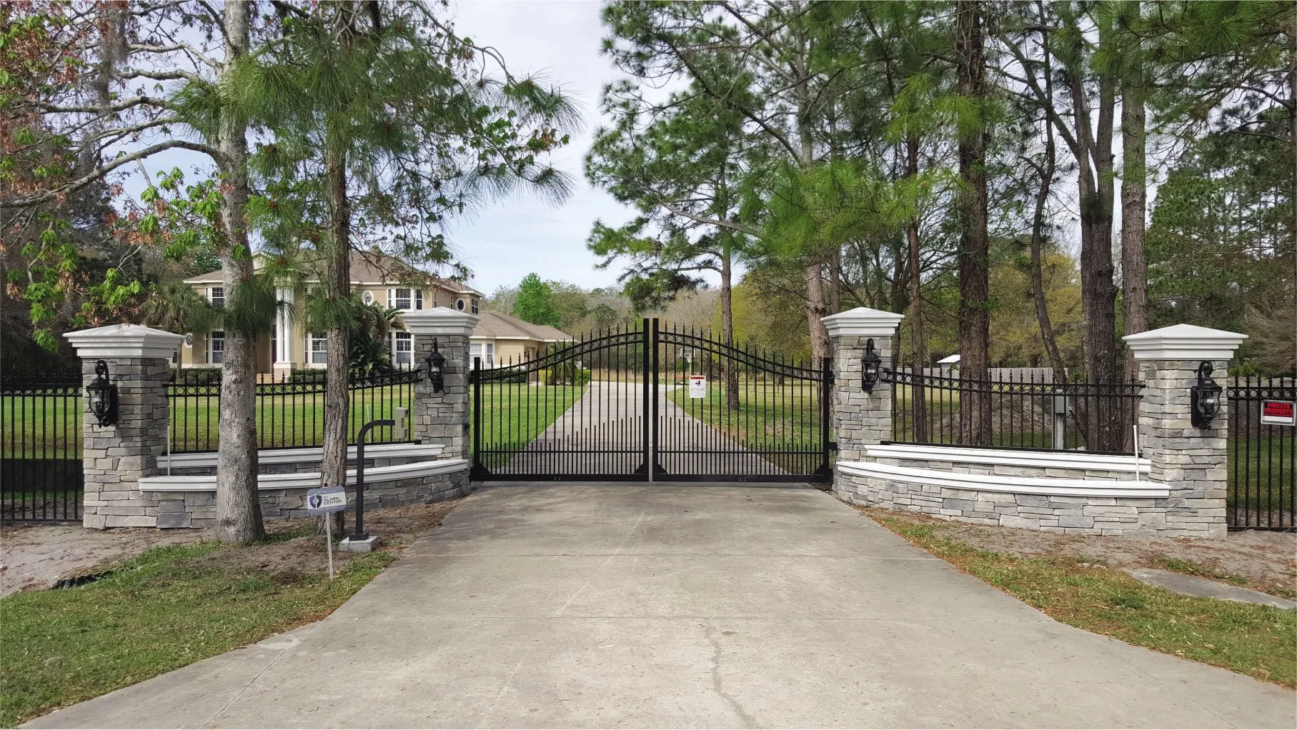 Black wrought iron gate with stone pillars, leading to a large house surrounded by trees and a well-maintained lawn.