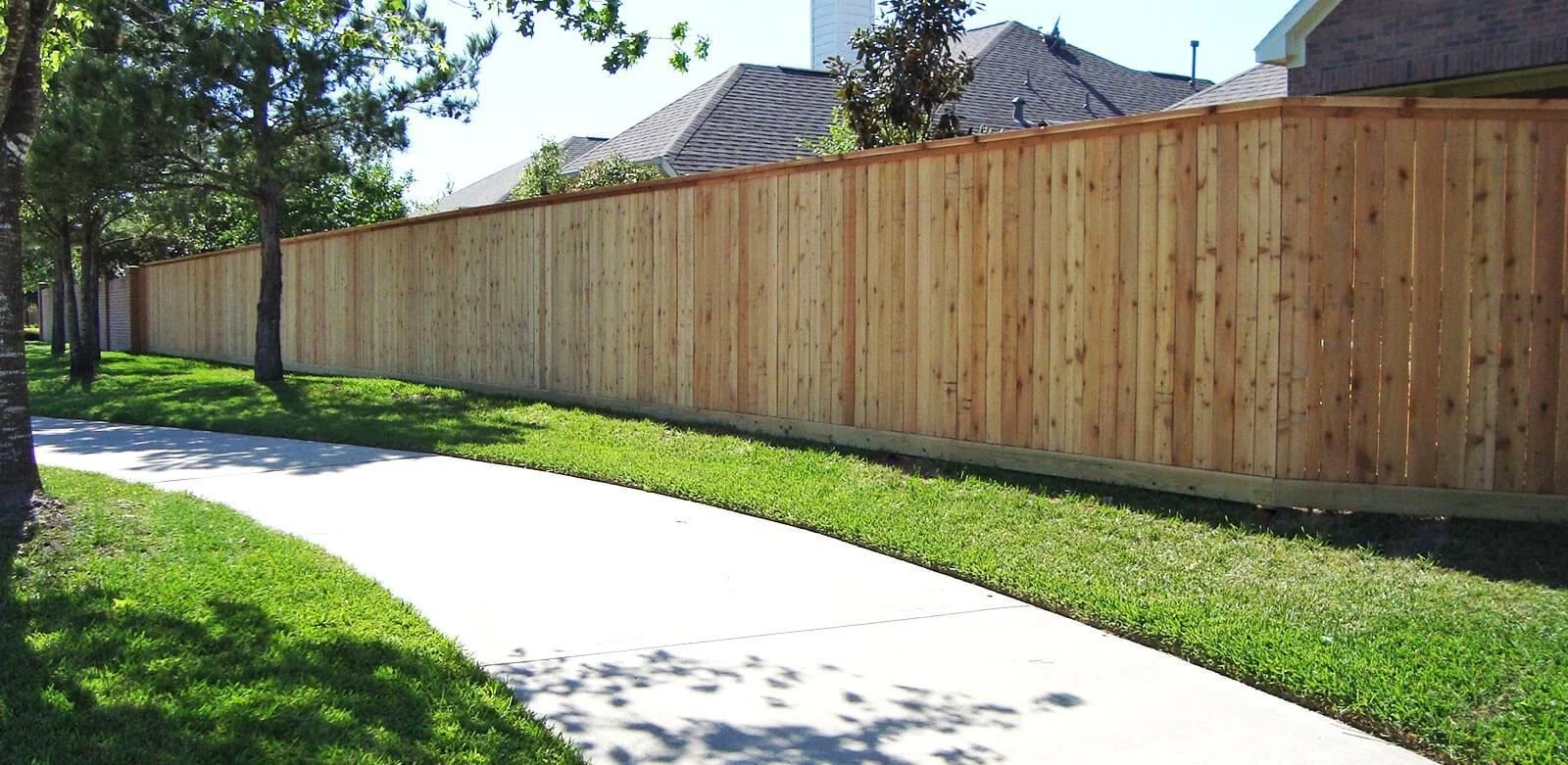 A tall wooden privacy fence runs alongside a sidewalk with a grassy verge, bordered by trees and houses in the background.