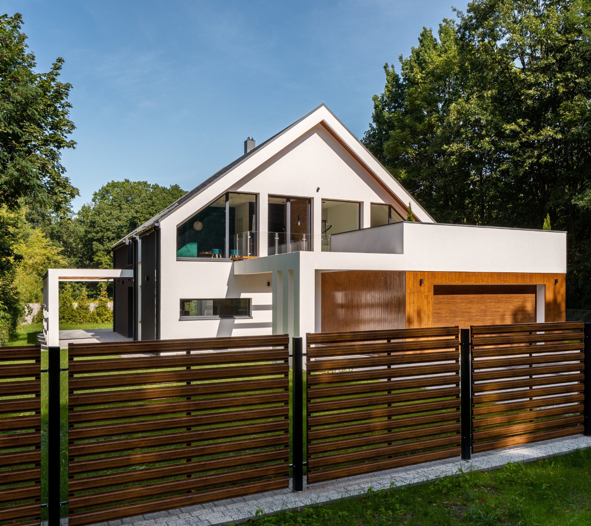 Modern white house with large glass windows and a wooden garage door, surrounded by trees and a wooden fence.
