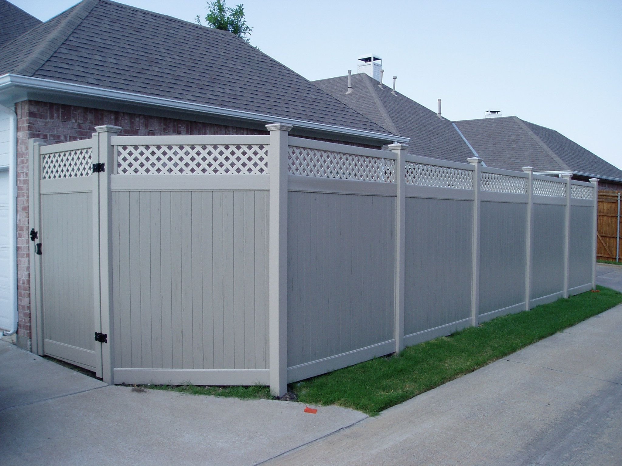 A vinyl privacy fence with lattice top section surrounds the backyard of a brick house with a shingled roof, a chimney, and vents. The yard has a concrete sidewalk and patches of grass.