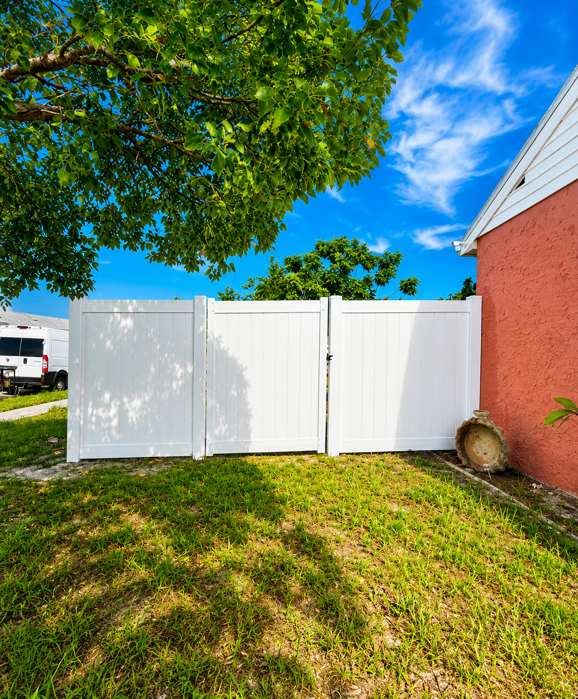 A white gate in front of a red stucco house, with a grassy yard and a large leafy tree overhead. There is a decorative garden ornament on the ground near the house wall, and a white van parked in the background under a clear blue sky with some clouds