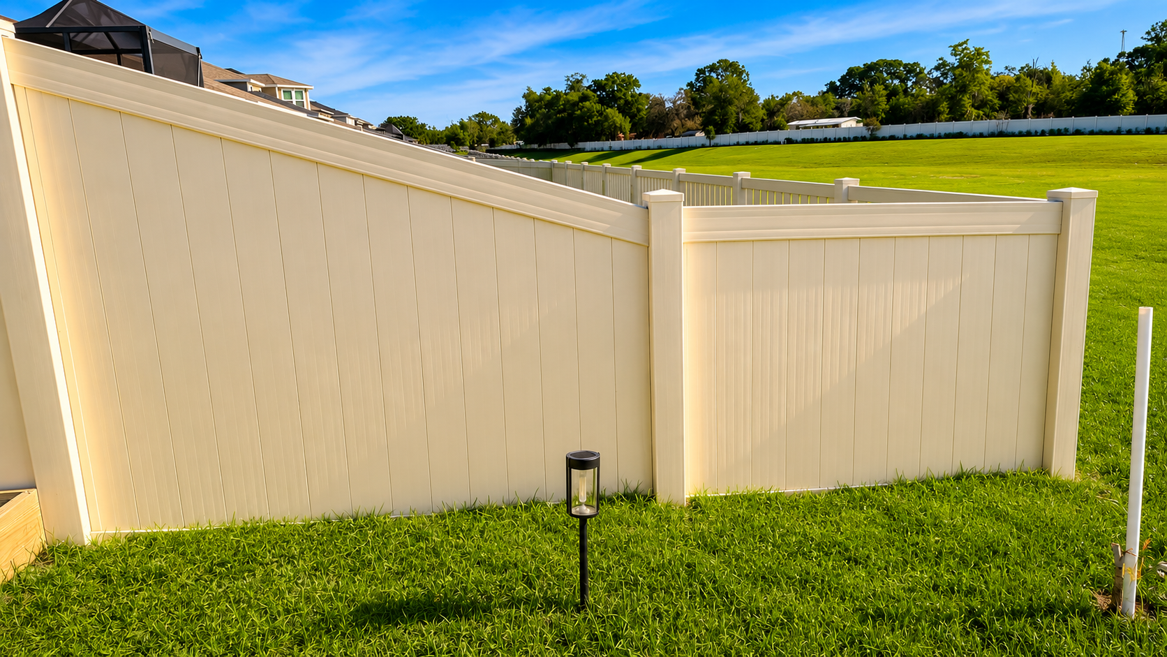 A beige vinyl privacy fence in a backyard, with a grassy yard and a small outdoor light in the foreground. In the background, there is a slope with a larger green field, trees, and houses under a clear blue sky.
