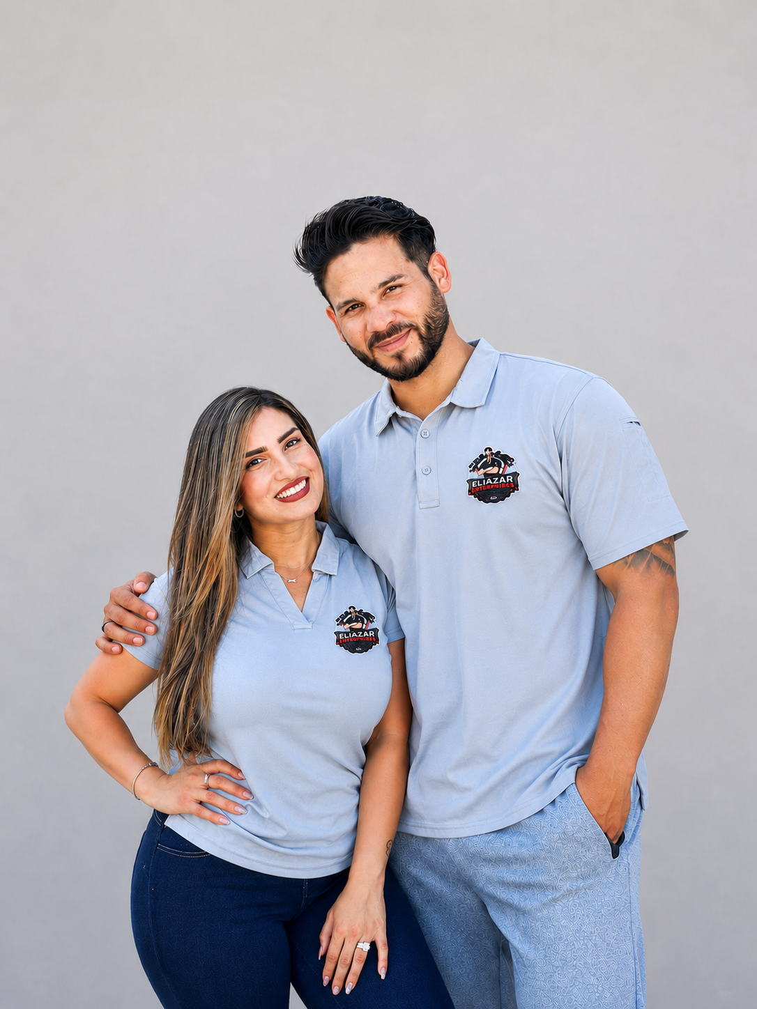 A smiling couple wearing matching light blue shirts with a logo, standing close together against a plain gray background.