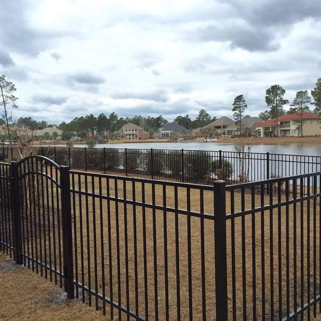 A backyard with a black metal fence, green grass, and a beige house with multiple windows and a small garden area.