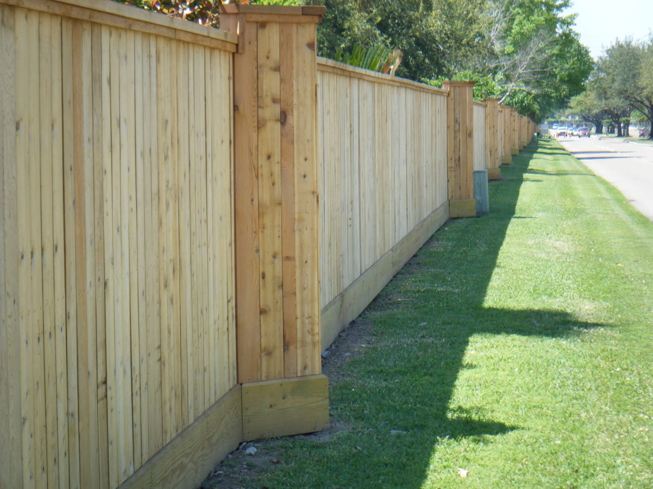 A long wooden privacy fence runs along a sidewalk, with neatly arranged vertical wooden planks and sturdy posts. Green grass covers the ground beside the fence, and trees with lush green foliage line the background, providing shade and a natural setting.