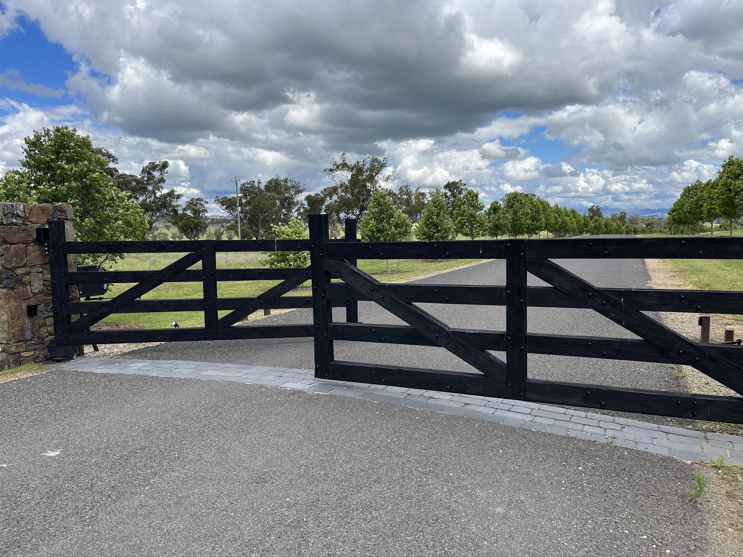 A wooden gate with a stone pillar on the left, opening to a gravel driveway lined with green trees under a partly cloudy sky.