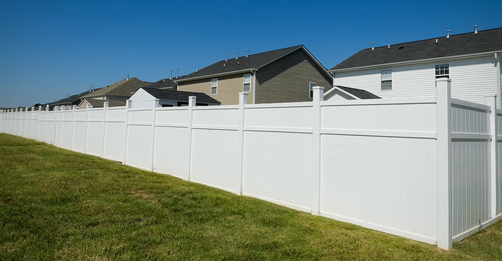 A white privacy fence runs alongside green grass with houses in the background, under a clear blue sky.