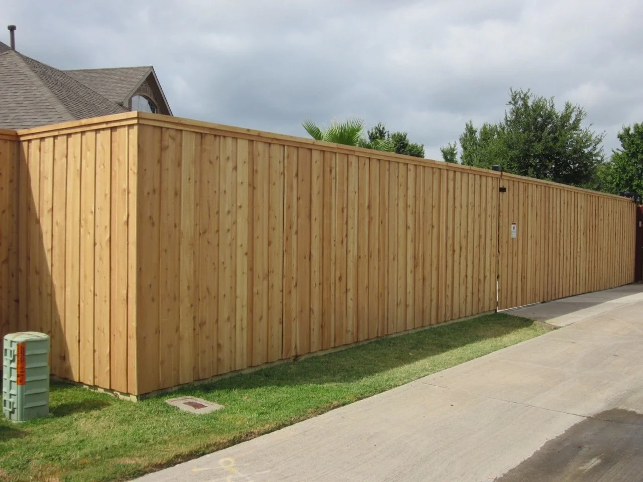 Long wooden privacy fence with a closed gate, adjacent sidewalk, and some greenery, under a partly cloudy sky.