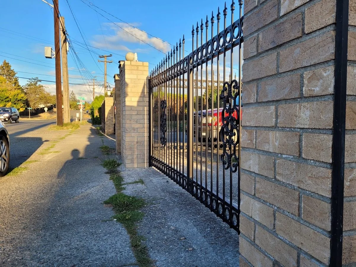 A black wrought iron gate attached to a brick wall, with cars and utility poles in the background on a sunny day.
