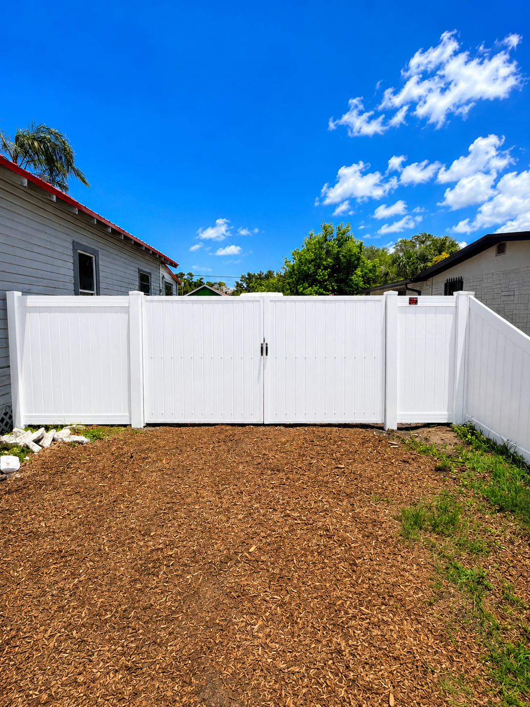 Backyard with white privacy fence, brown dirt ground, neighboring houses, green trees, blue sky, with clouds.