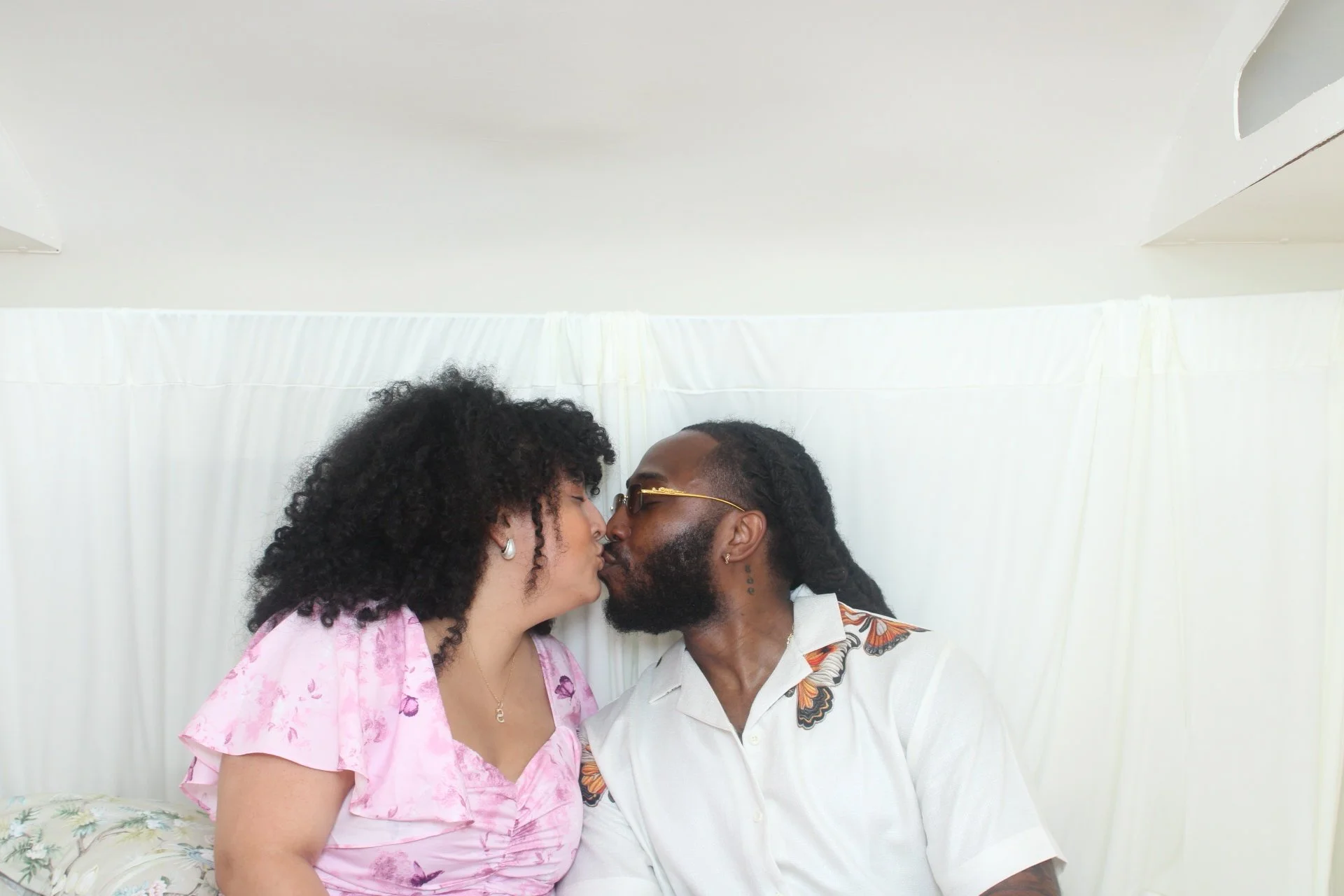 A couple is sharing a kiss in front of a white curtain. The woman is wearing a pink blouse with purple patterns and has curly black hair. The man has glasses, a beard, and is wearing a white shirt with a butterfly pattern.