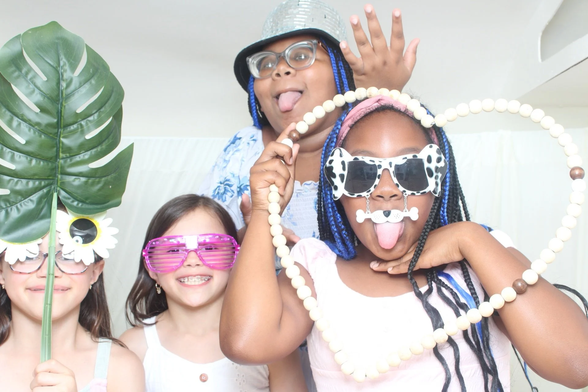 Group of children wearing colorful sunglasses, holding props like a large leaf with sunflower decorations, and making playful faces while dressed up in fun accessories at a celebration.