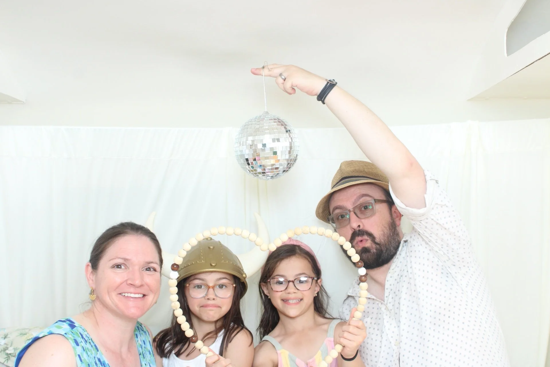 Group of five people, two adults and three children, at a party, posing for a selfie, with a disco ball hanging from the ceiling in the background. The man is holding a makeup mirror shaped like a heart, and a girl is wearing a Viking helmet.