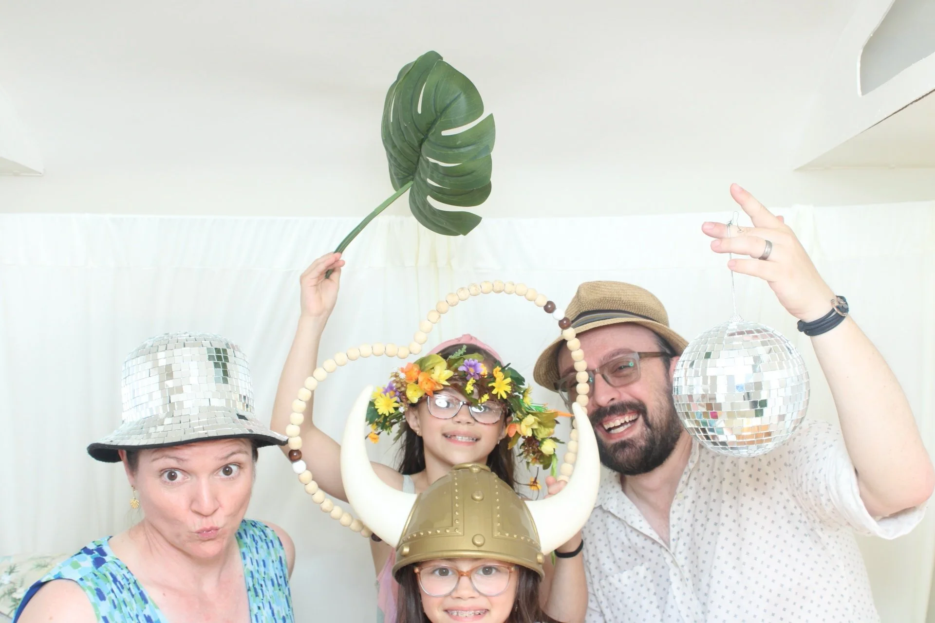 Group of people celebrating, wearing costumes like a witch hat, adorned with flowers, and holding disco balls and decorative items, smiling and posing indoors.