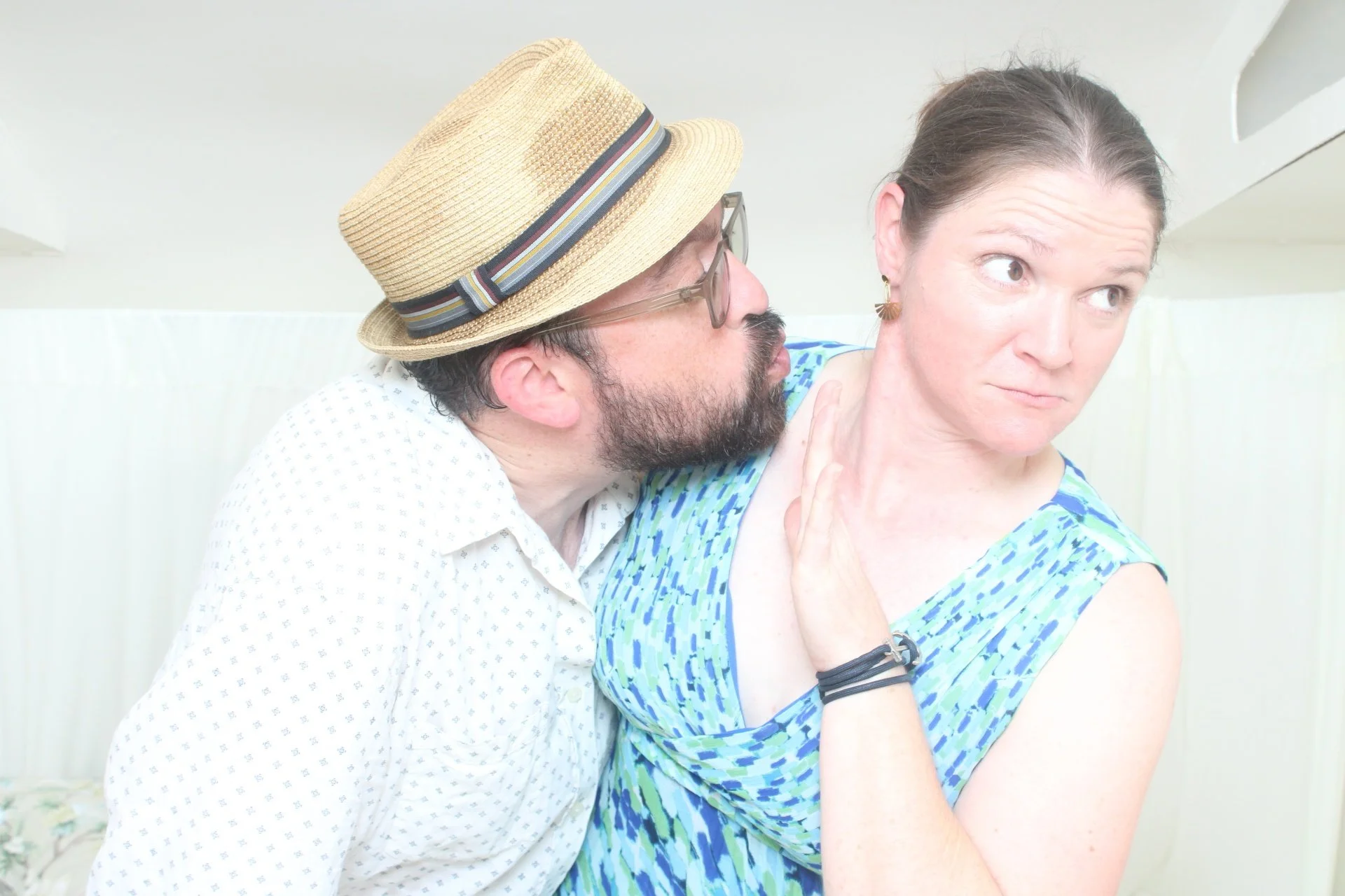 A man wearing a straw hat and glasses leans in close to a woman in a blue patterned dress, who looks away with a skeptical expression.