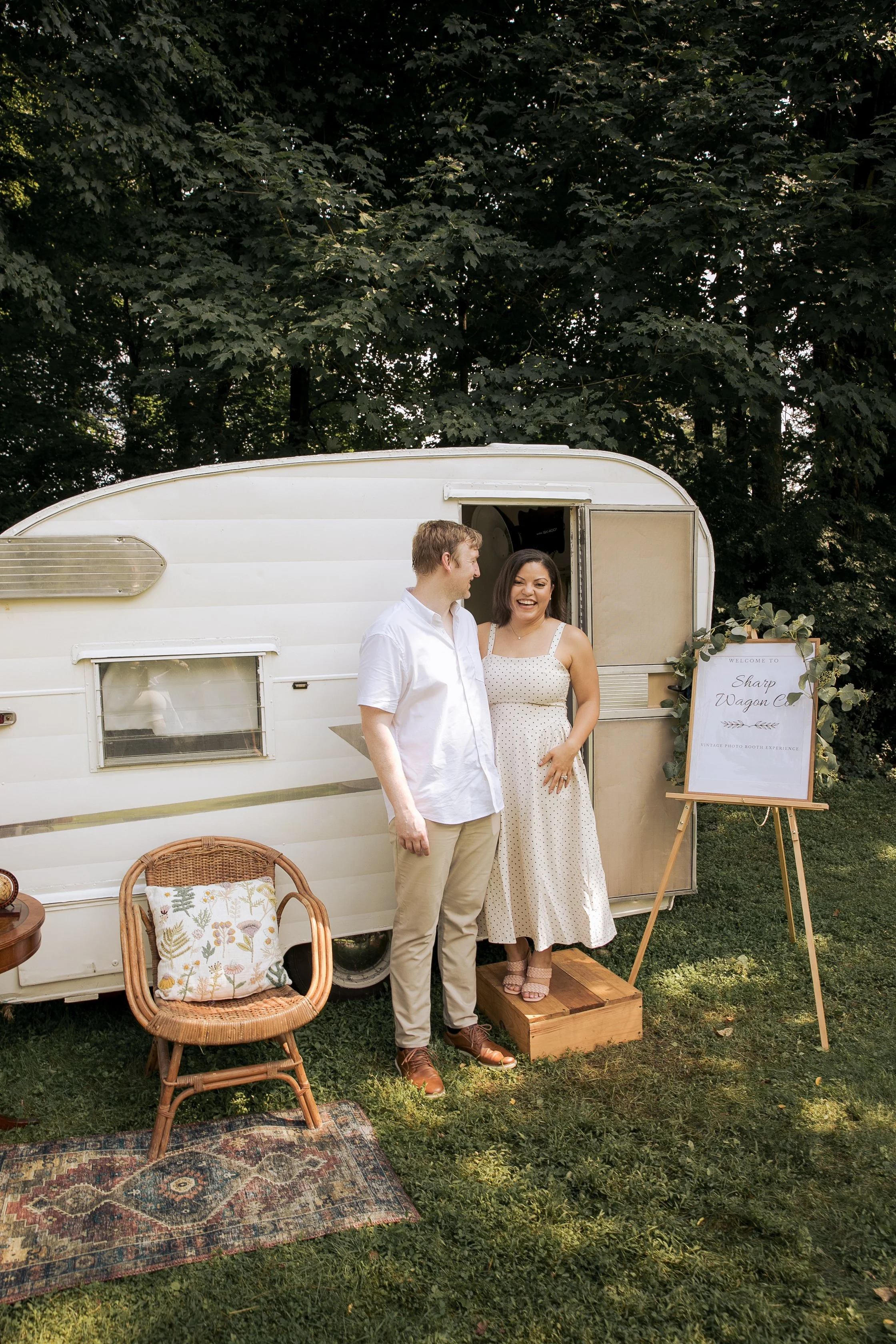 A couple standing in front of a vintage camper trailer at an outdoor event, engaging with each other and smiling. There is a sign on an easel nearby and outdoor furniture including a wicker chair and a colorful rug on the grass.