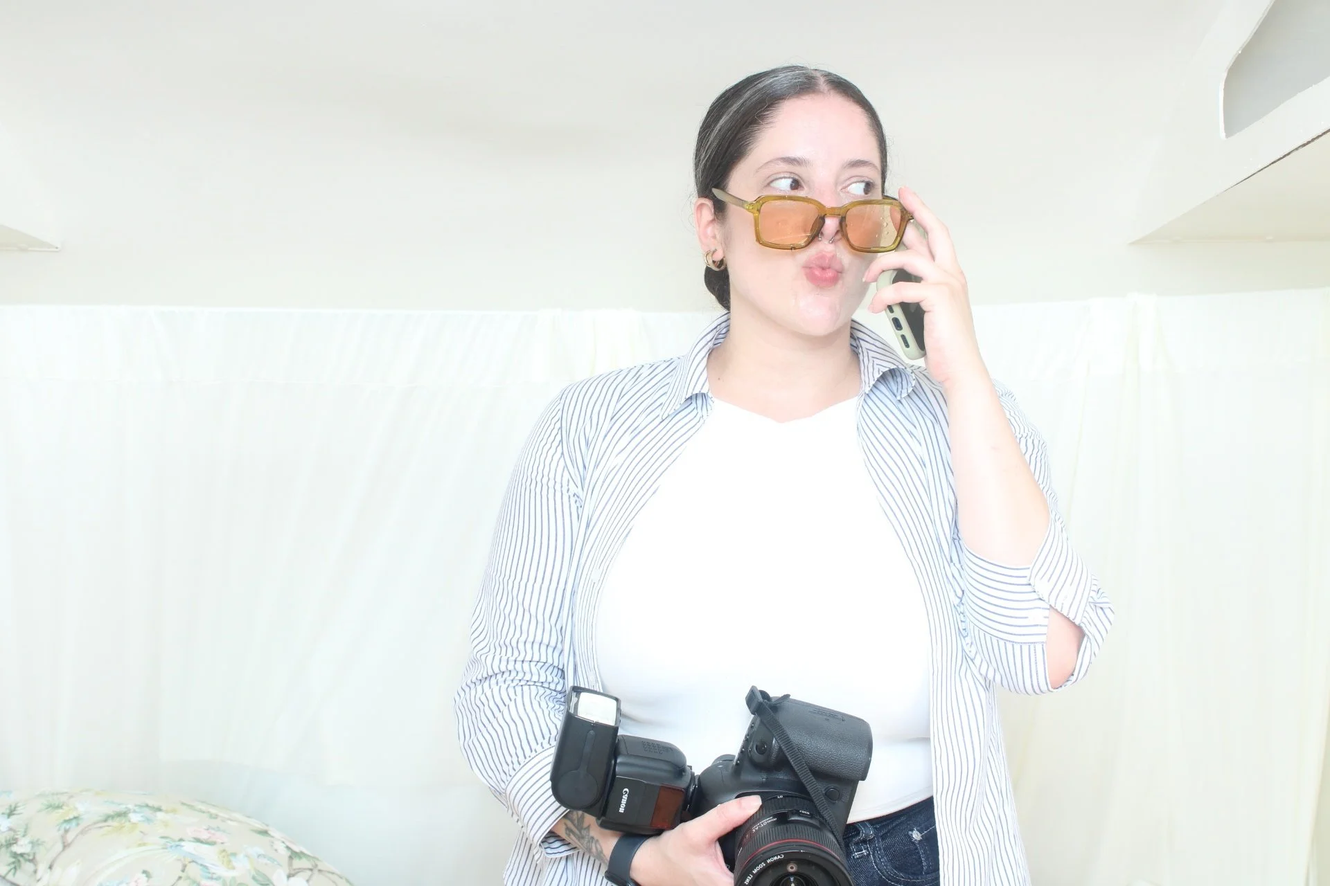 Woman with dark hair, wearing sunglasses and a striped shirt, holding a camera and talking on a cellphone in a bright room.