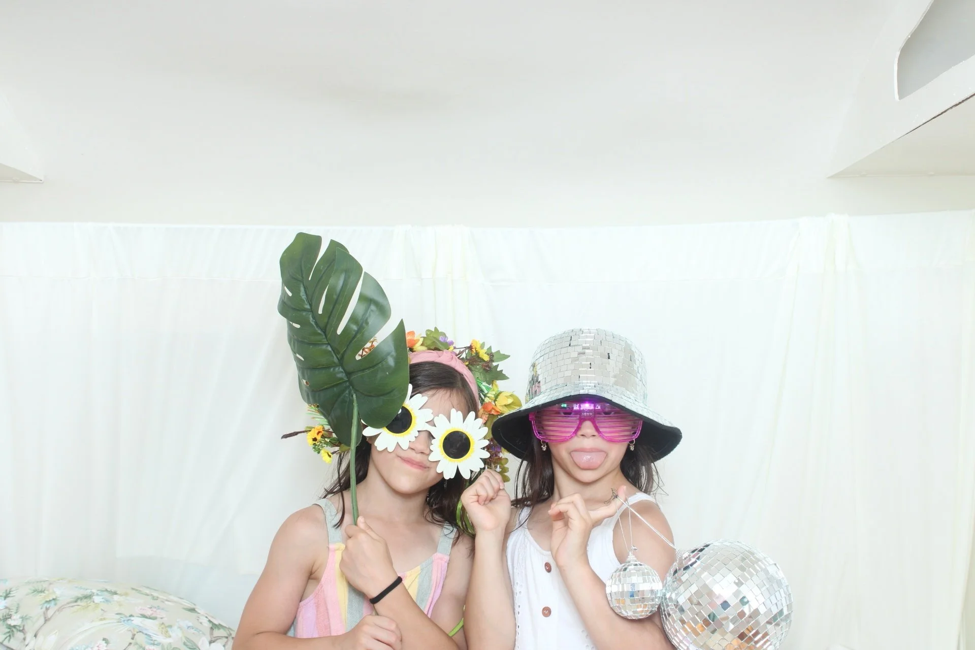 Two women wearing fun accessories: one with large sunglasses and a flower crown, and the other with a silver disco ball hat and pink shutter shades, posing with balloons in a photo booth with a white backdrop.