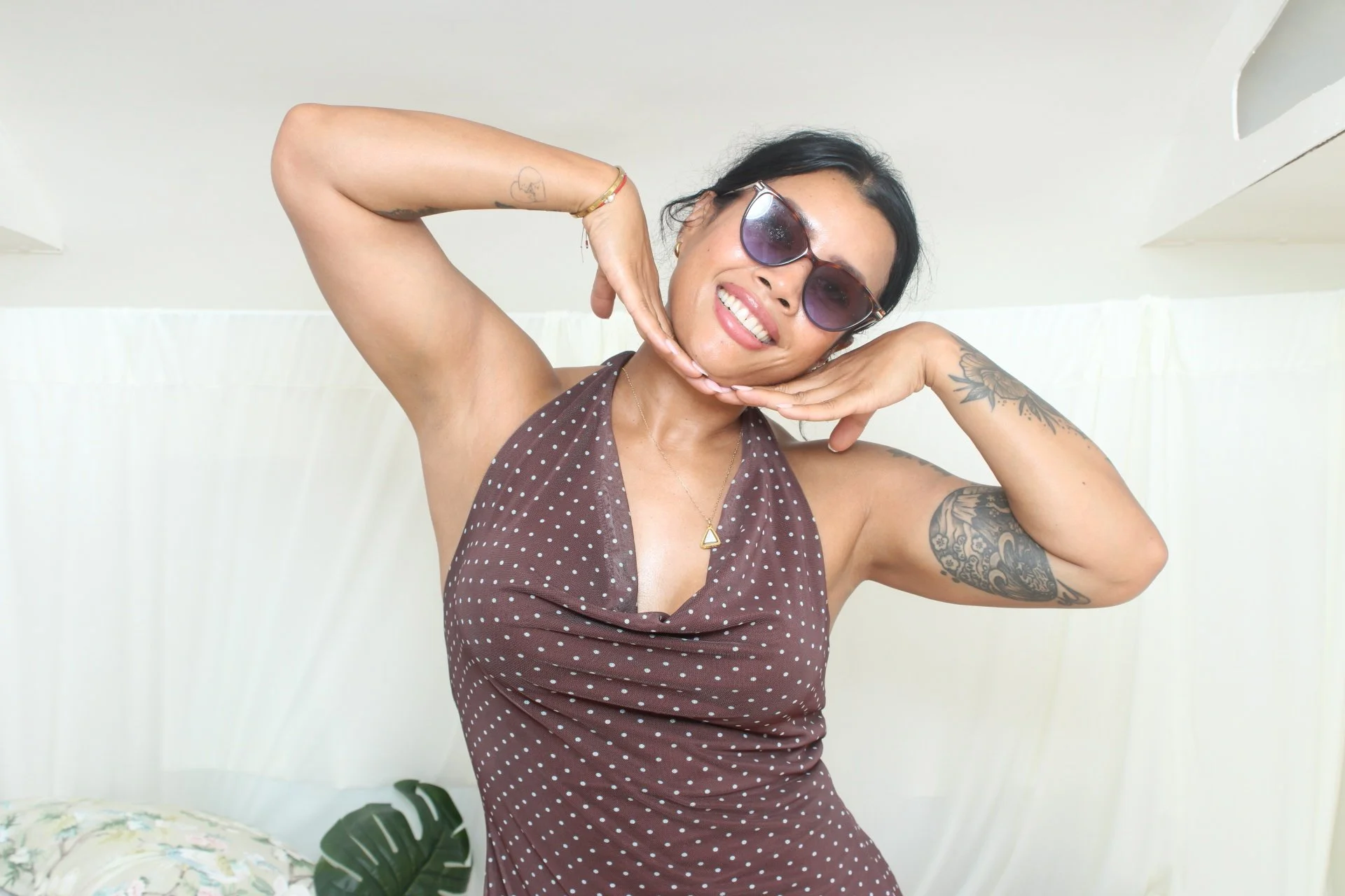 Woman with dark hair wearing sunglasses and a brown polka dot dress, smiling and posing with her hands under her chin in front of a white background.