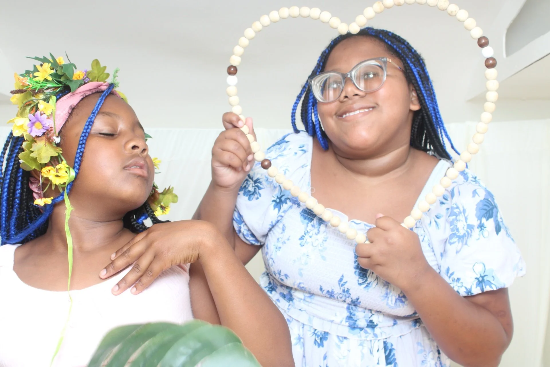 Two women with blue braided hair and glasses smiling, one holding a beaded heart-shaped frame and the other wearing a flower crown.