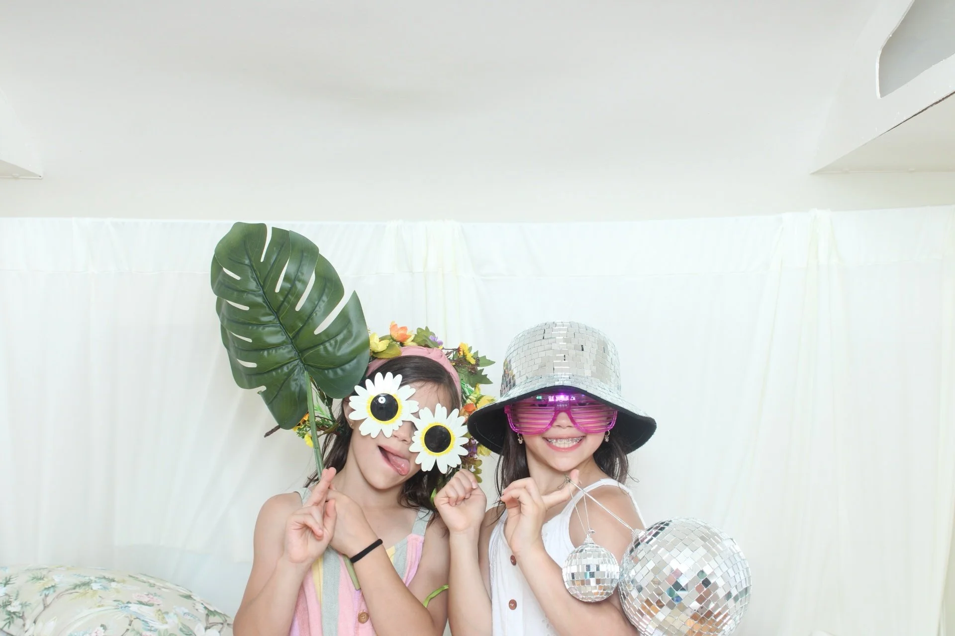 Two young women smiling and posing with playful accessories in a photo booth. One wears oversized sunglasses, a shiny disco ball hat, and a flower headband with large white daisies. The other wears large pink shutter shades, a straw hat, and a flower