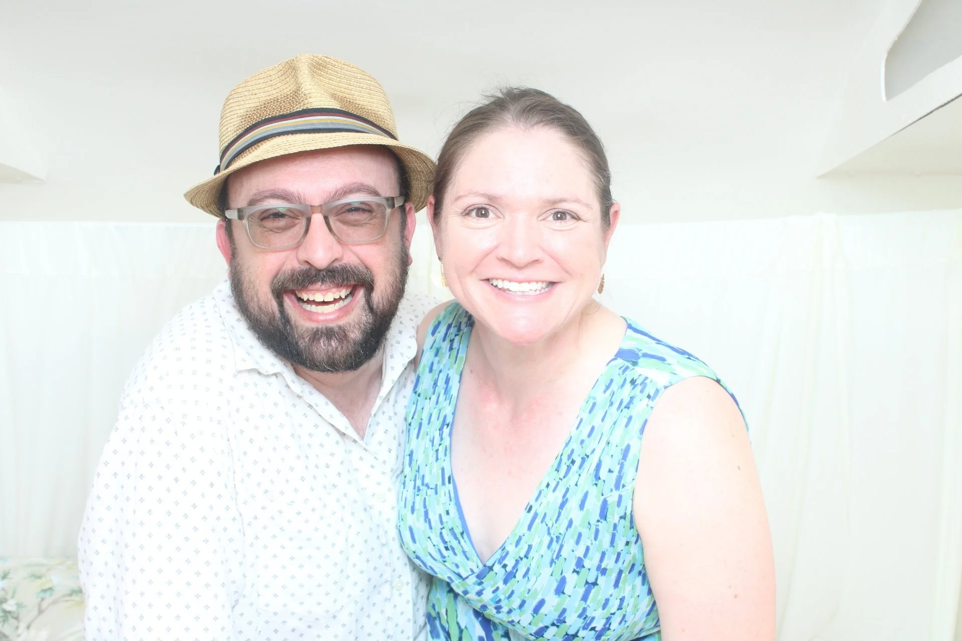 A smiling couple, a man with a beard wearing glasses and a straw hat, and a woman with brown hair in a blue patterned dress, posing closely together indoors in a light-colored room.