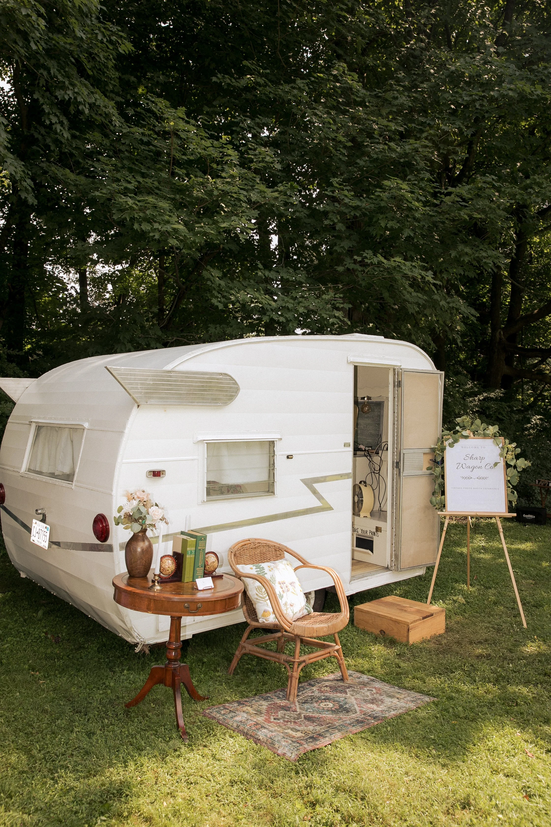 A vintage trailer used as a photo booth with a sign that reads 'Sharp Wagon Co.' on an easel, set up outdoors with chairs, a small table with books and flowers, a patterned rug, and surrounded by trees.