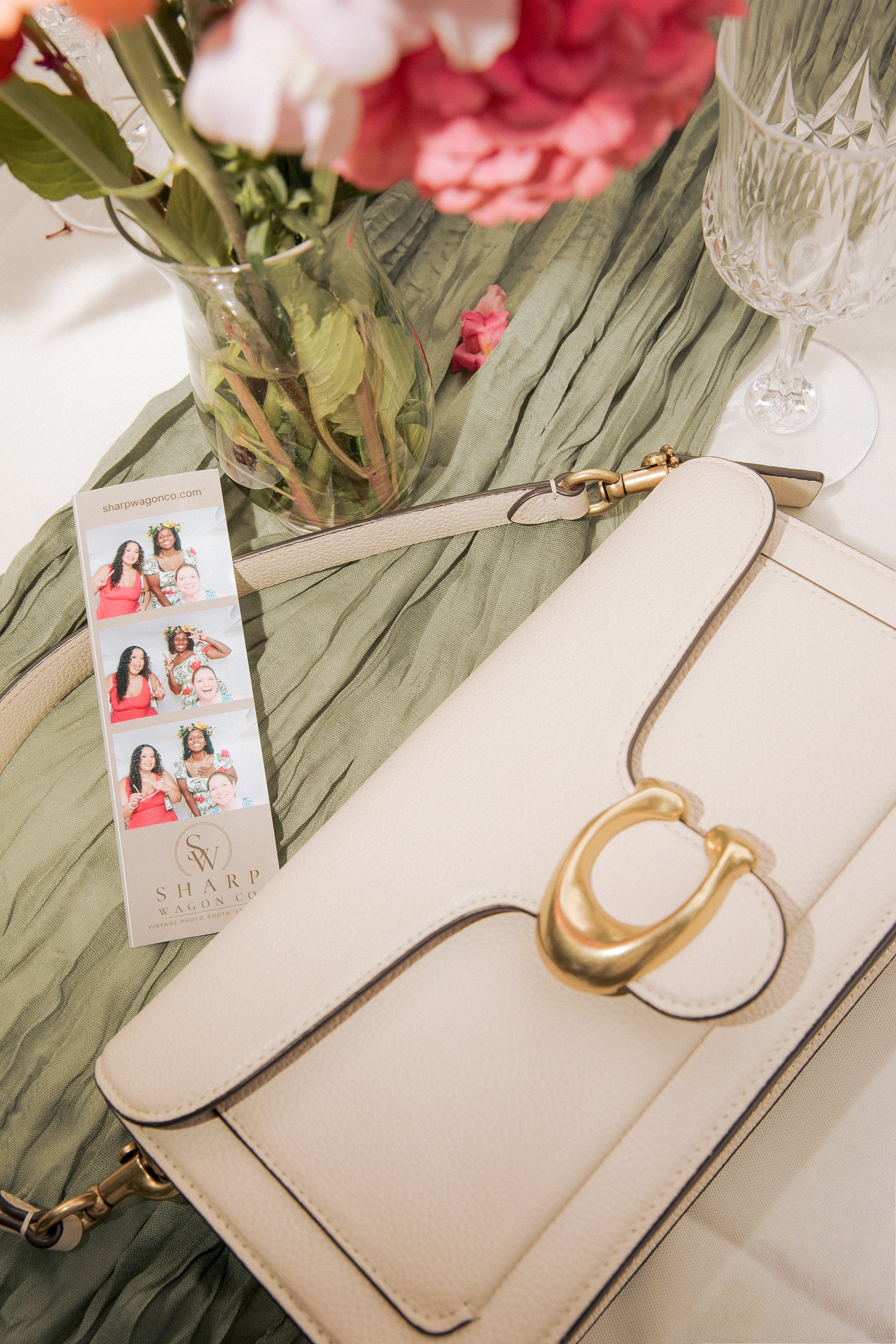 Close-up of a beige jewelry box with gold jewelry, a palm leaf-patterned glass, and a flower in a vase on a table with a green fabric runner, along with a photo booth strip and a business card from Sharp Wagon Co.