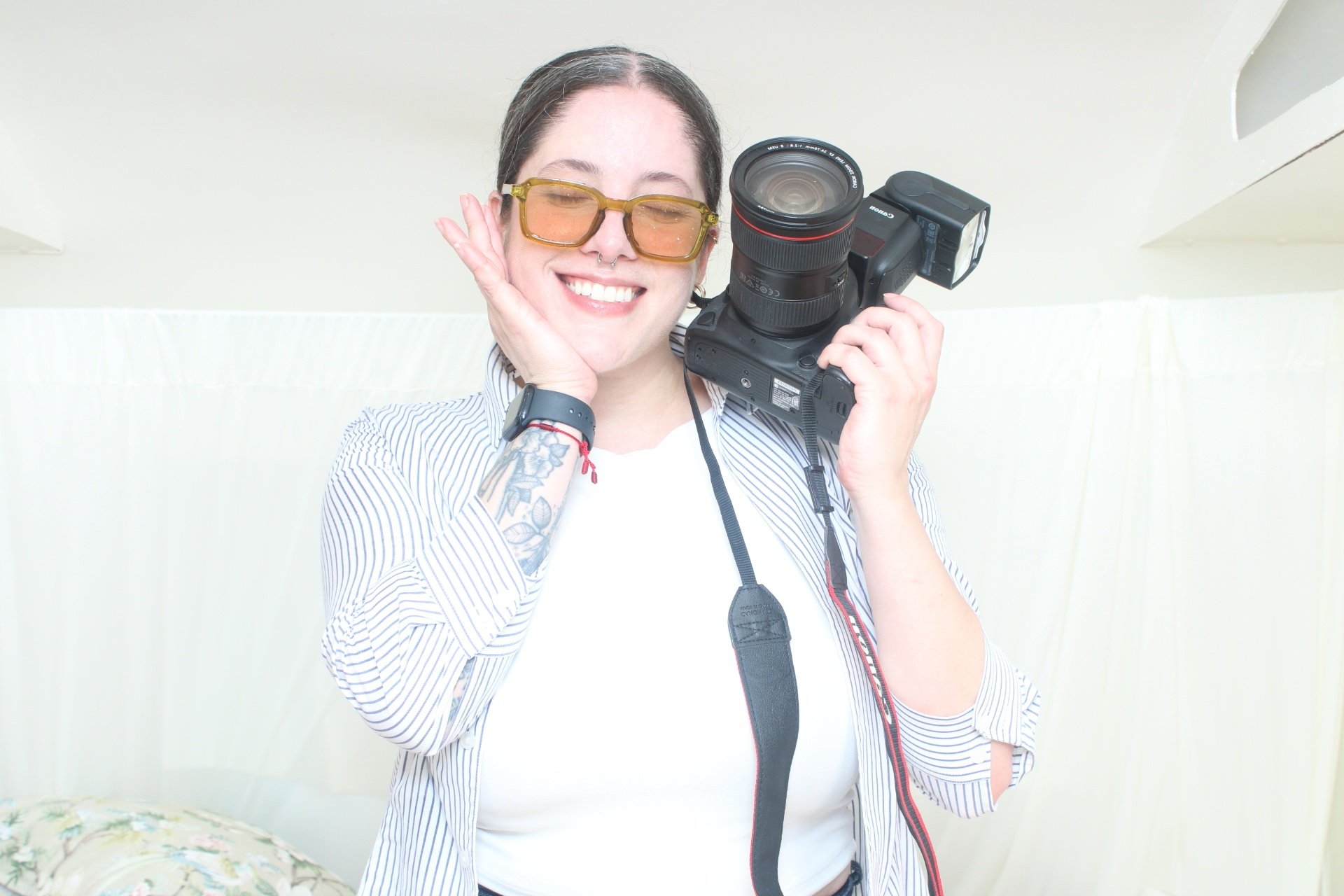 A smiling woman with tattoos on her arm holding a camera, wearing sunglasses and a striped shirt.