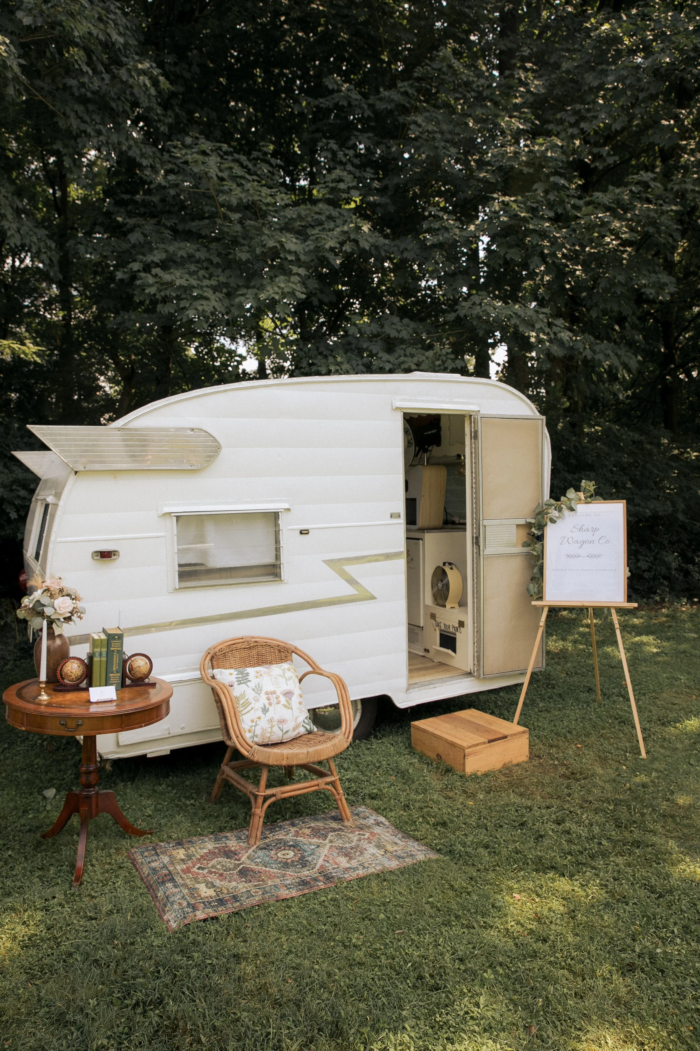 A vintage camper converted into a small outdoor cafe or vendor booth, decorated with plants and vintage items, set on green grass with trees in the background.