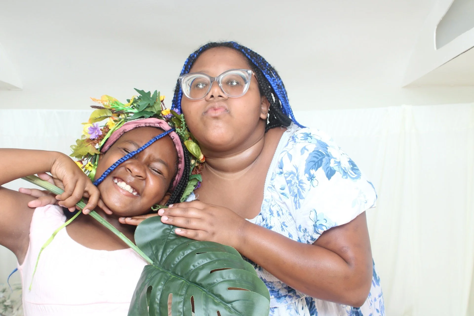 A girl with a floral crown and a woman with blue braided hair and glasses posing inside a bright room.