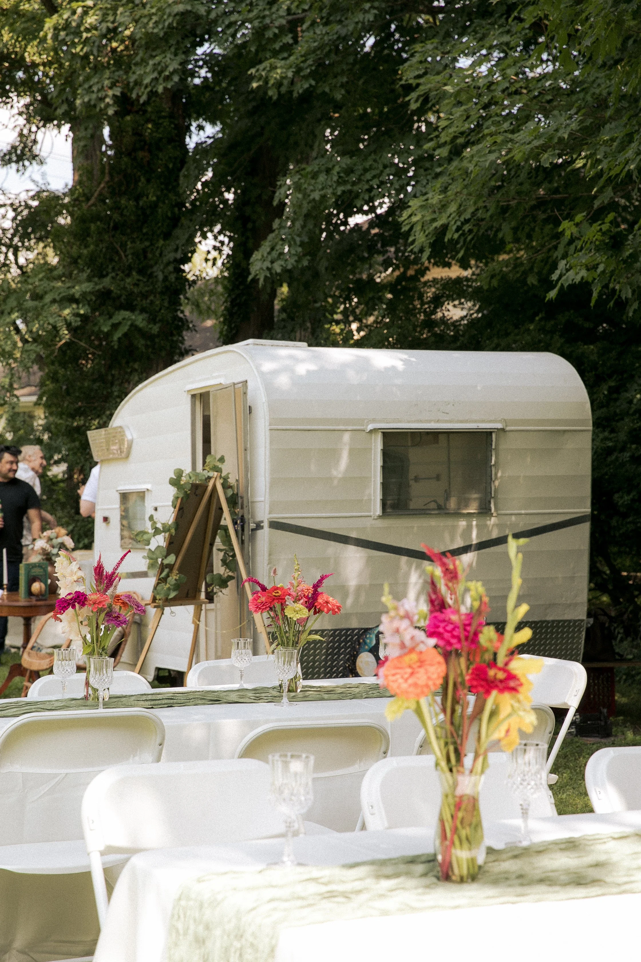 Outdoor gathering with decorated tables, floral centerpieces, and a vintage camper trailer surrounded by trees.