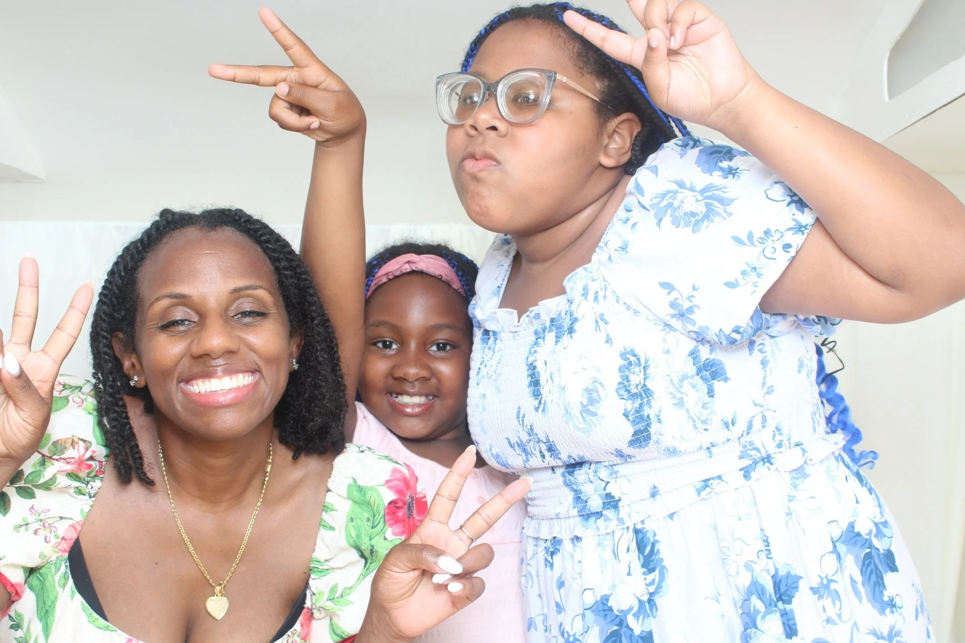 Three women and a girl smiling and making peace signs with their hands, wearing floral dresses, indoors.