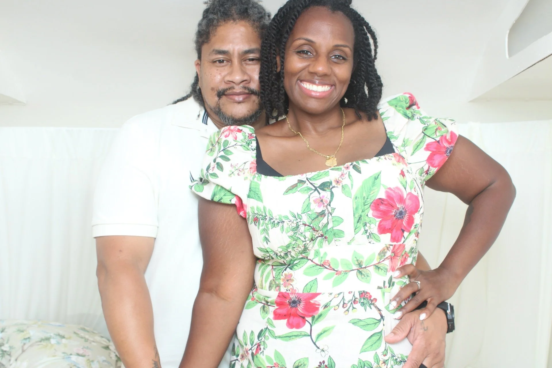 A man and woman standing together, smiling, in a bright room with white walls and a shelf. The woman wears a floral dress, and the man wears a white shirt with black stripes on the collar. The woman has dark, styled hair and the man has dreadlocks wi