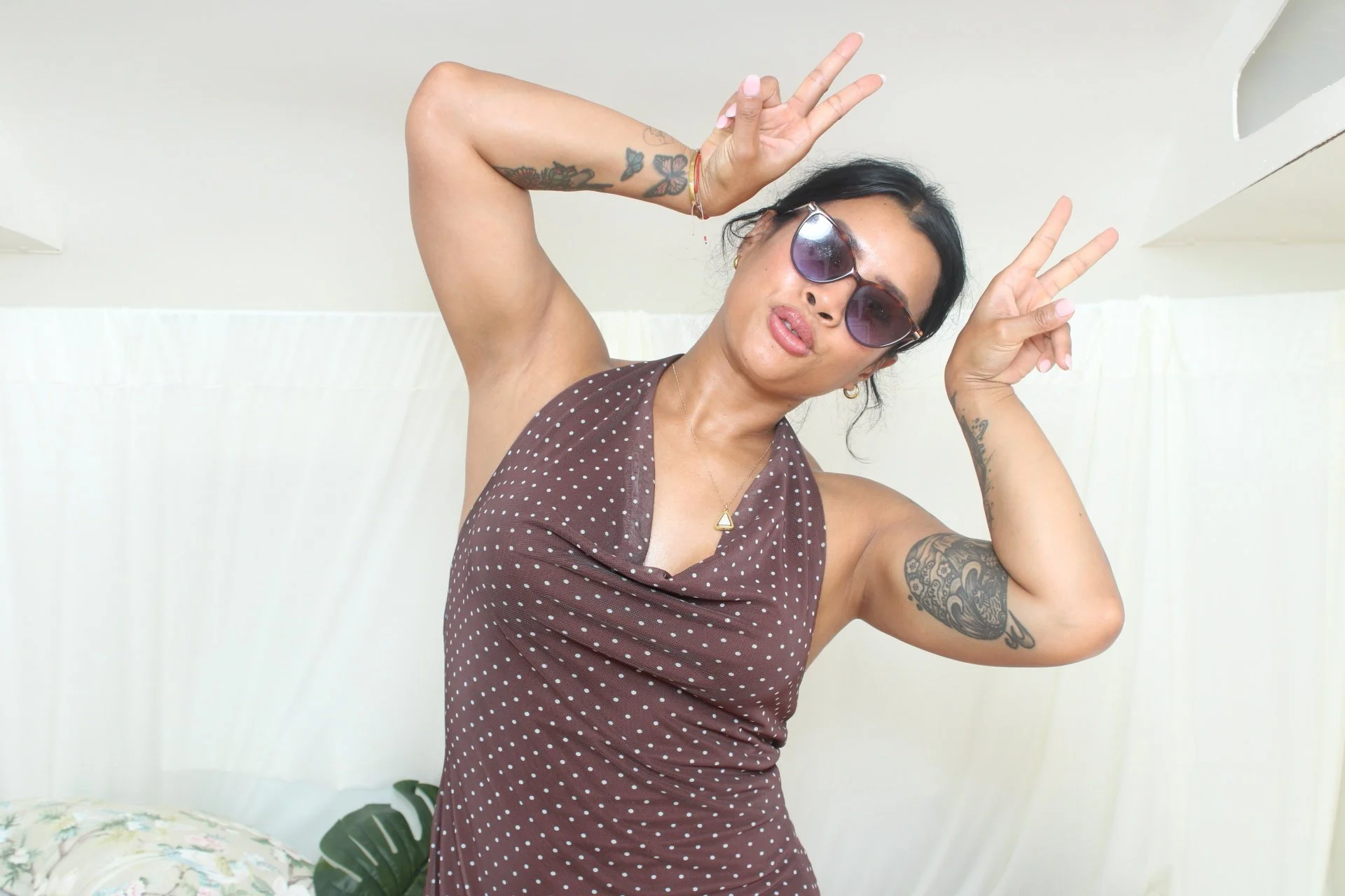 A woman with dark hair, wearing sunglasses and a brown polka-dot dress, making peace signs with both hands and posing for the camera in a room with a white curtain background.