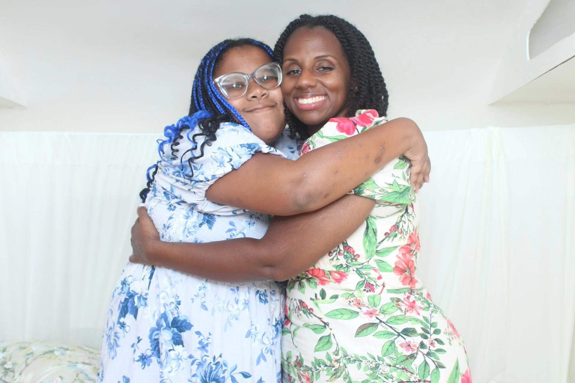 Two women hugging and smiling at the camera, one wearing glasses and a blue and white floral dress, the other wearing a colorful floral dress.